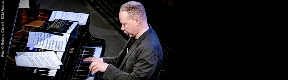 Ein Mann spielt konzentriert Klavier, umgeben von Notenblättern, die auf dem Flügel liegen.