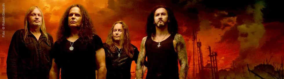 Vier Männer in dunkler Kleidung stehen vor einem apokalyptischen, rot-orange leuchtenden Hintergrund.