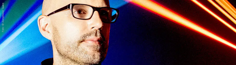 Ein Mann mit Brille vor bunten Lichtstrahlen, die einen energischen, futuristischen Hintergrund bilden.