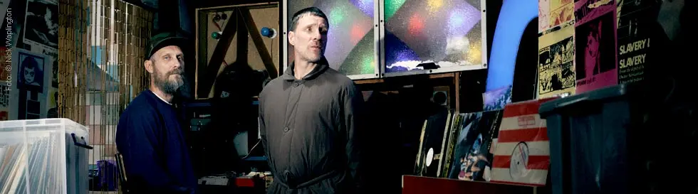 Zwei Männer stehen in einem Raum voller Platten und elektronischer Geräte. Der Raum wirkt dunkel und kreativ gestaltet.