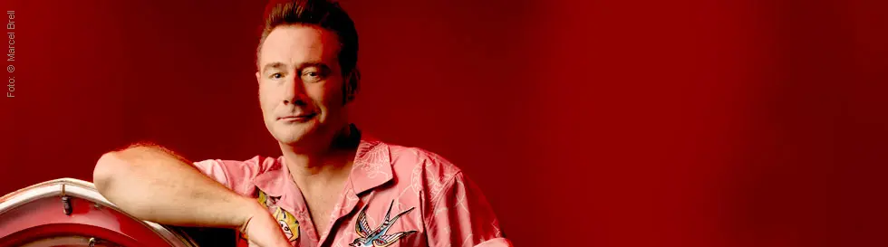 Ein Mann in einem pinken Hemd mit Schwalbenmuster lehnt sich auf einer Jukebox, roter Hintergrund.