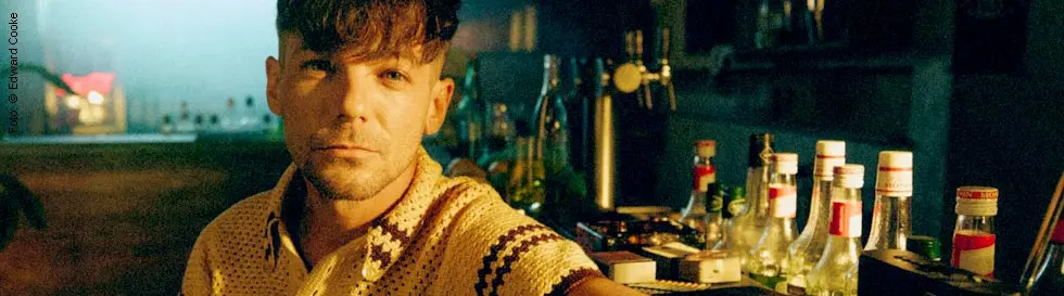 Mann in gestreiftem Shirt an Bar, Flaschen hinter ihm, gedämpftes Licht, entspannte Atmosphäre, Pflanzen rechts.