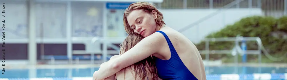 Eine junge Frau in einem blauen Badeanzug umarmt ein Kind in einem geblümten Badeanzug. Sie sitzen am Rand eines Schwimmbeckens.