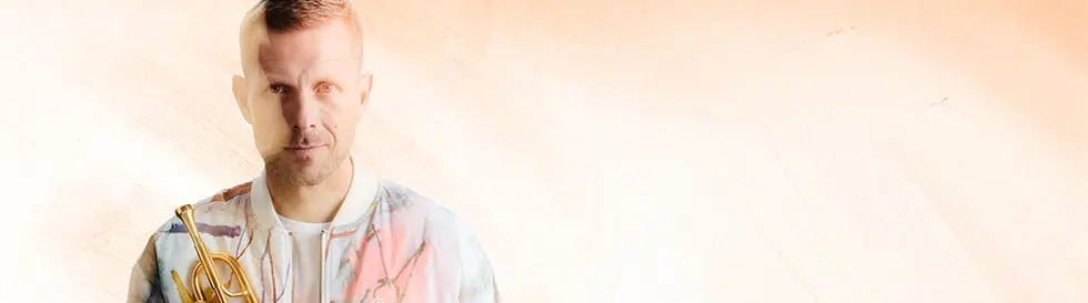 Ein Mann hält eine Trompete, trägt eine bunt gemusterte Jacke und steht vor einem hellen, unscharfen Hintergrund.