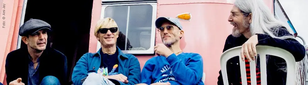 Vier Menschen sitzen entspannt auf Plastikstühlen vor einem rosafarbenen Gebäude.