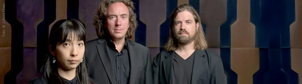 Drei Personen in schwarzer Kleidung vor einer geometrischen, gemusterten Wand.