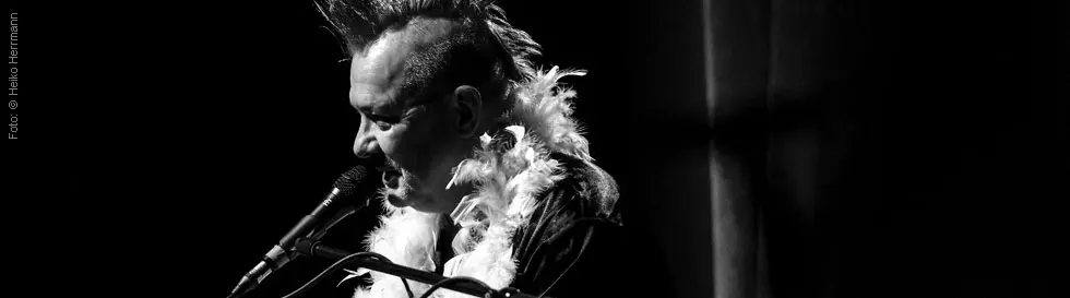 Person mit Irokesenfrisur am Mikrofon und Federboa, schwarz-weiß.