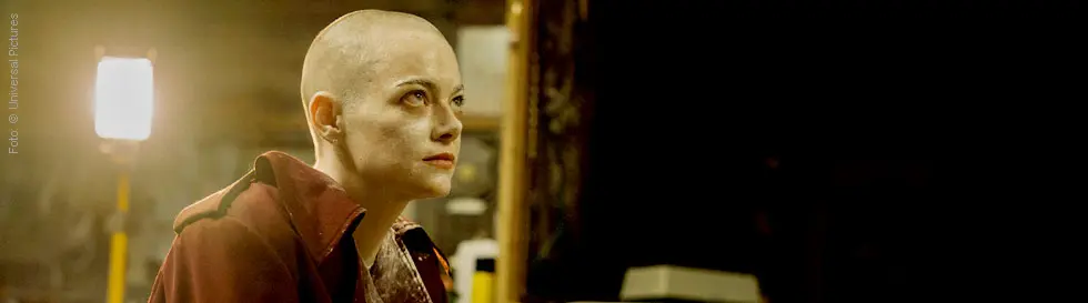 Eine Person mit Glatze in einem roten Mantel sitzt in einem spärlich beleuchteten Raum und schaut nachdenklich.