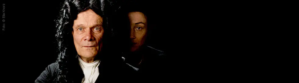 Zwei Gesichter vor dunklem Hintergrund, älterer Mann vorn mit schwarzer Perücke, Frau im Schatten dahinter.