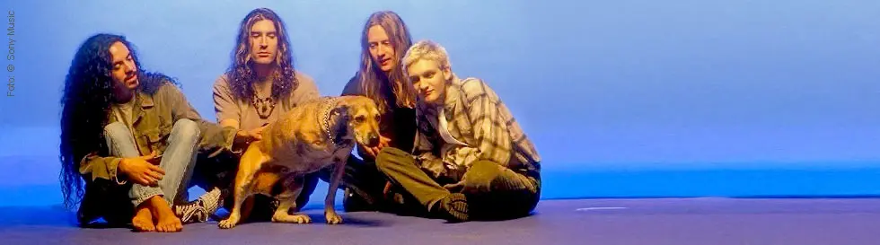 Vier Menschen sitzen nebeneinander auf dem Boden vor einem blauen Hintergrund, mit einem Hund in der Mitte.