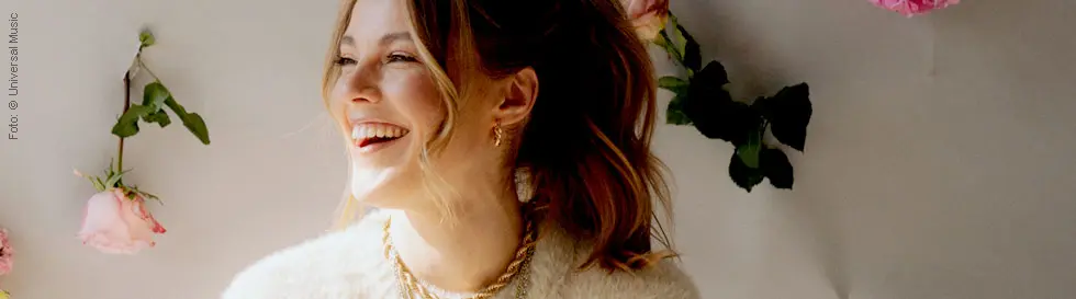 Eine lächelnde Frau mit blonden Haaren, umgeben von hängenden rosa Blumen. Sie trägt einen flauschigen Pullover.