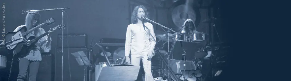 Ein Musiker im weißen Anzug singt auf einer Bühne; links spielt jemand Gitarre; Schlagzeuger im Hintergrund.