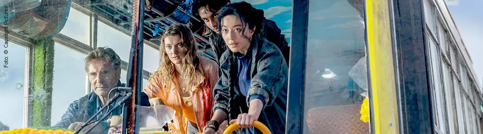 Vier Personen in einem Bus nach Kathmandu, eine Person am Lenkrad. Blumengirlanden schmücken den Innenraum, die Frontscheibe ist zerschossen.