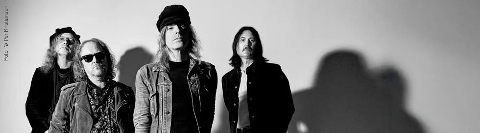 Vier Männer in schwarz-weißer Fotografie, tragen Hüte und Sonnenbrillen, stehen lässig nebeneinander.