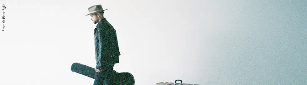 Ein Mann mit Hut trägt ein Gitarrenkoffer, geht an zwei alten Koffern vorbei. Minimalistischer Hintergrund.