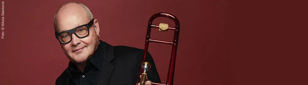 Ein Mann mit Brille und Glatze hält eine Posaune vor einem dunkelroten Hintergrund.