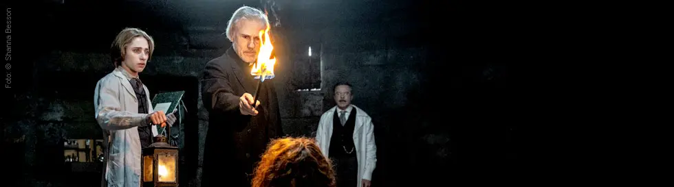 Dunkle Szene mit drei Männern, einer hält ein brennendes Kreuz vor einer sitzenden Person.