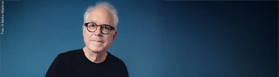 Ein älterer Mann mit weißem Haar und Brille sitzt vor einem blauen Hintergrund. Er trägt ein schwarzes Oberteil.