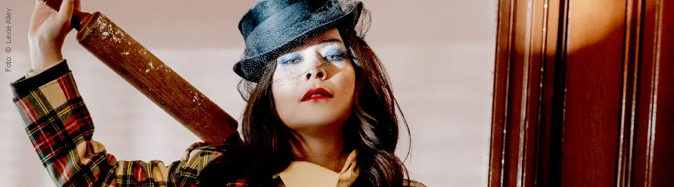 Eine Frau in bunter Jacke, Hut mit Netz, blauem Lidschatten, rotem Lippenstift, hält einen Nudelholz.