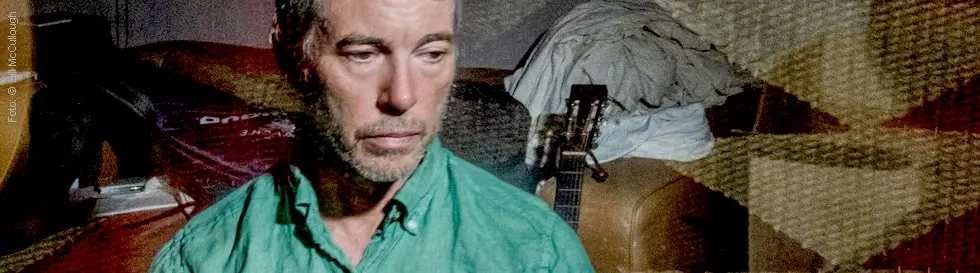 Ein nachdenklicher Mann in einem grünen Hemd sitzt vor einer Sofaecke, im Hintergrund eine Gitarre und Kleidung.