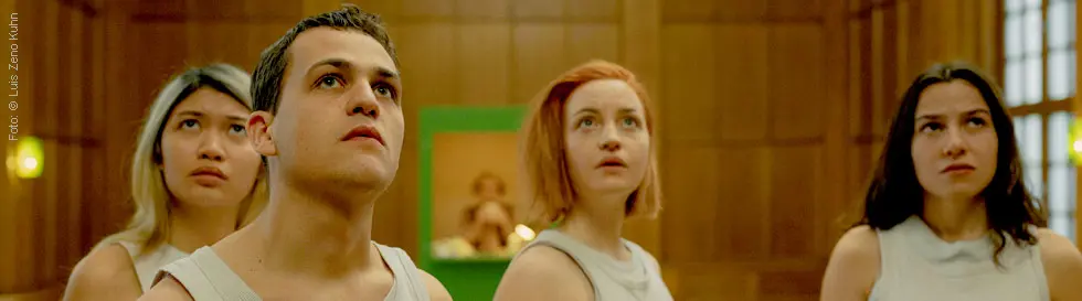 Vier junge Menschen in weißen Outfits blicken nach oben in einem holzgetäfelten Raum.