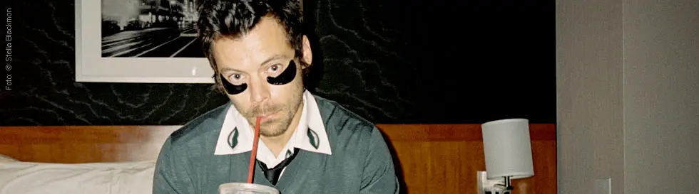 Person mit dunklen Augenstreifen sitzt auf einem Bett und trinkt aus einem Strohhalm.