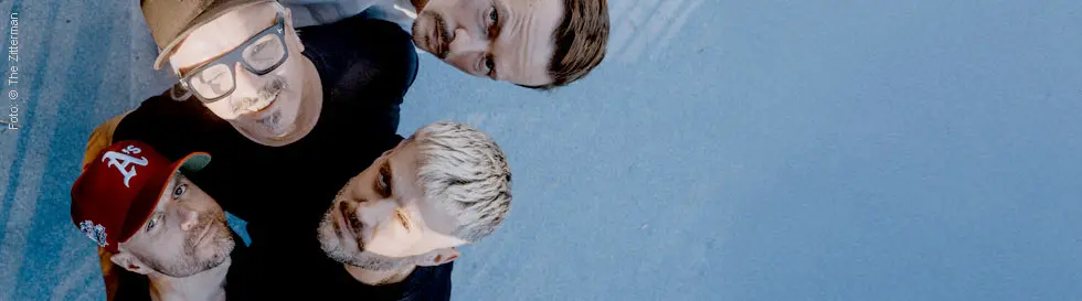 Vier Männer schauen nach oben, einer trägt eine rote Kappe, ein anderer hat blonde Haare und einer eine Brille.