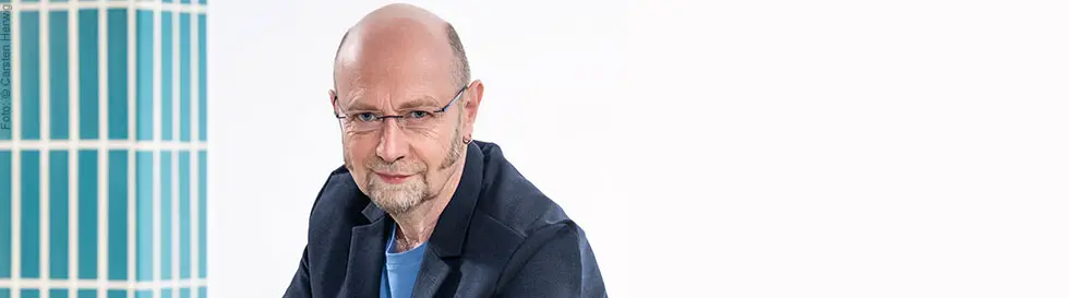 Ein Mann mit Brille und Bart sitzt lächelnd im Vordergrund, im Hintergrund vertikale blaue Streifen.