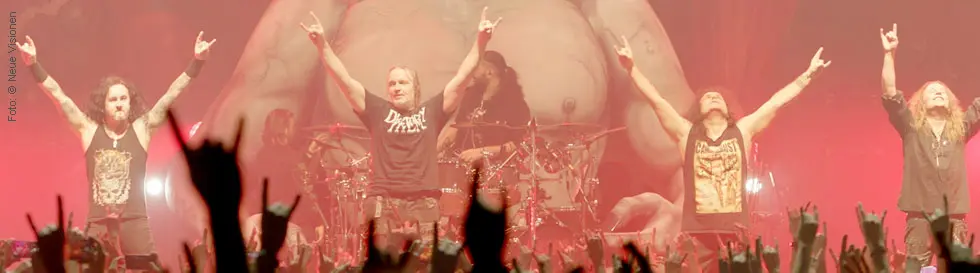 Fünf Männer stehen mit erhobenen Händen auf einer Bühne. Davor jubelt eine Menge und zeigt das Rockhandzeichen.