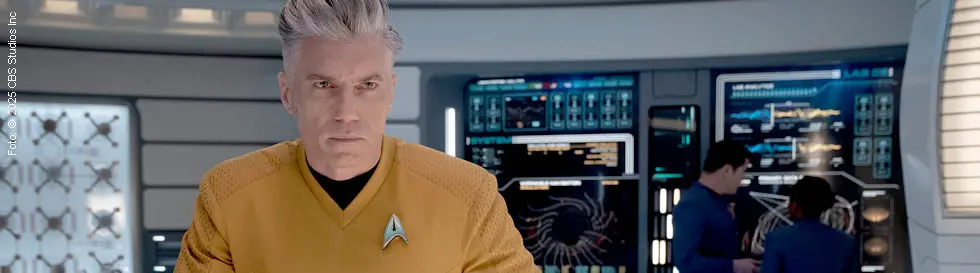 Ein Mann in gelber Uniform steht ernst in futuristischem Raum, im Hintergrund digitale Bildschirme.