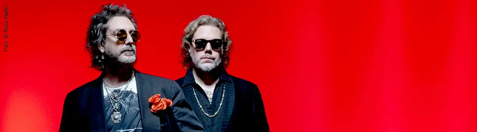 Zwei Männer mit Sonnenbrillen vor rotem Hintergrund, beide in dunklen Anzügen, einer mit orangefarbenem Einstecktuch.