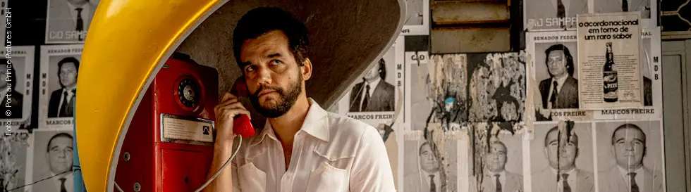 Ein Mann telefoniert in einer gelben Telefonzelle neben Plakaten, die das Wort "Marcos Freire" zeigen.