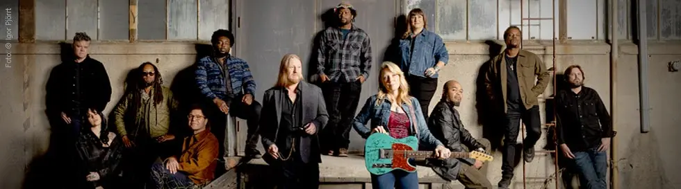 Zwölf Personen in lässiger Kleidung posieren vor einem Industriebau, eine Frau in der mitte hält eine türkise E-Gitarre.