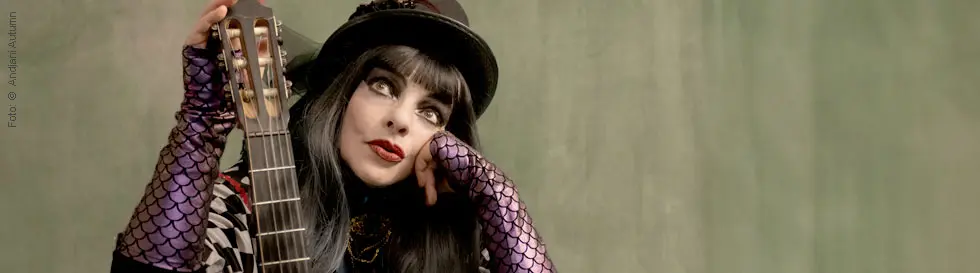 Eine Frau mit dunklem Haar, buntem Make-up und Hut hält eine Gitarre, trägt ein kariertes Outfit und fingerlose Netzhandschuhe.