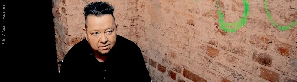 Eine Person mit aufgerichteten dunklen Haaren sitzt an einem Tisch vor einer Ziegelwand und blickt nachdenklich.