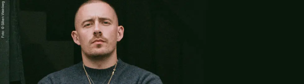 Person mit kurzem Haar, grauem Pullover, Ketten, schaut ernst in die Kamera.