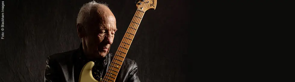 Eine ältere Person hält eine E-Gitarre und trägt eine schwarze Lederjacke.