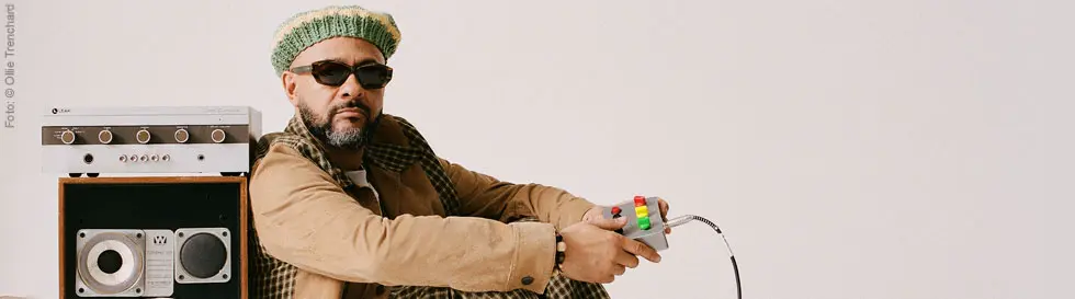 Ein Mann mit grün-beiger Mütze sitzt lässig neben Lautsprechern und hält einen Controller. Foto: © Ollie Trenchard. 