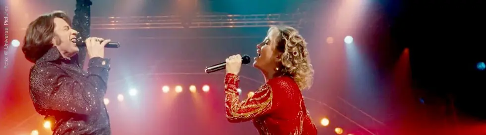 Links steht ein Mann und eine Frau in glamouröser Kleidung singen mit Mikrofonen auf einer beleuchteten Bühne.