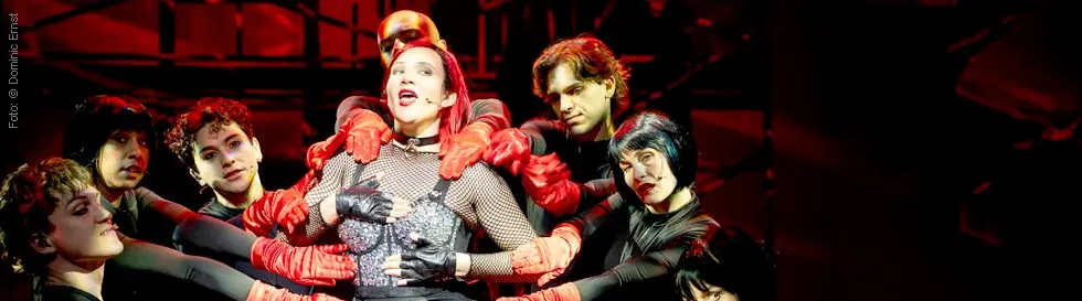 Menschen in schwarzer Kleidung mit roten Handschuhen posieren dynamisch, während eine zentrale Person im Netzoberteil singt.