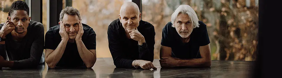 Vier Männer liegen nebeneinander auf einem Boden, mit den Ellbogen aufgestützt. Sie schauen direkt zur Kamera.