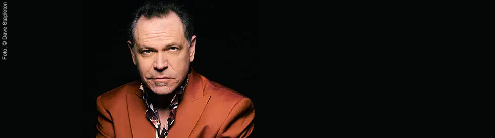 Ein Mann mit einem entschlossenen Blick trägt ein orangefarbenes Sakko und ein gemustertes Hemd.