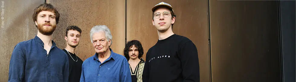 Fünf Männer in Freizeitkleidung posieren vor einer braunen Wand. Einer trägt eine Brille und Mütze.