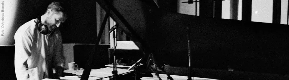 Ein Mann mit Kopfhörern steht an einem Flügel, umgeben von Noten und einem Mikrofon.