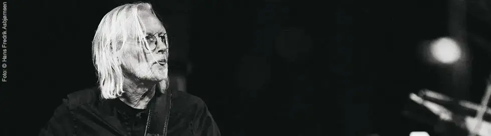 Ein Gitarrist mit langen Haaren spielt konzentriert. Er trägt eine Brille und dunkle Kleidung.