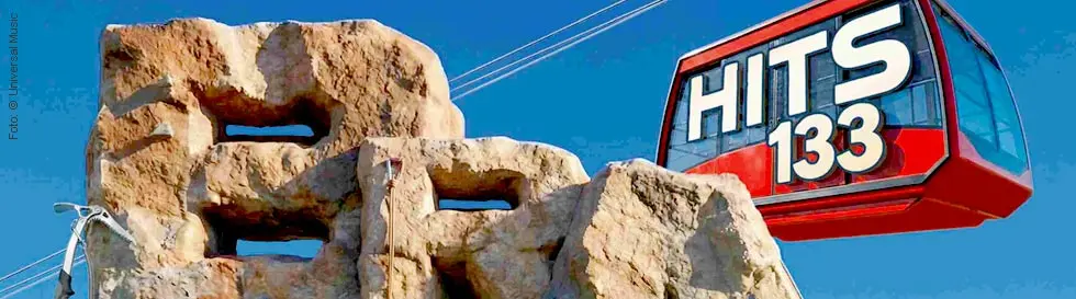 Große Steinskulptur mit den Buchstaben "BRAVO". Luftseilbahnwagen mit der Aufschrift "HITS 133" vor blauem Himmel.