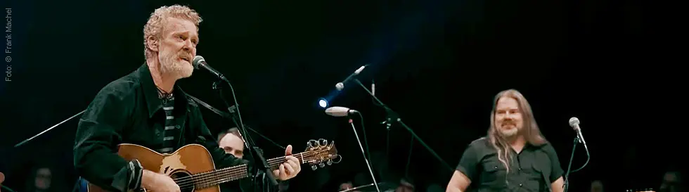 Ein Mann singt und spielt Gitarre, daneben steht ein weiterer Mann mit Bass, beide auf einer Bühne mit Mikrofonen.