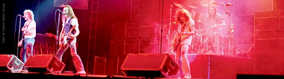 Vier Musiker auf einer Bühne mit ihren Instrumenten, umgeben von buntem Licht und Nebel.