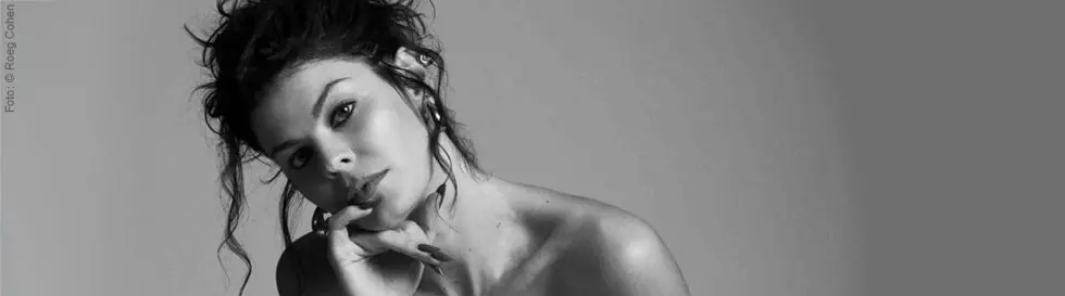 Eine Frau mit lockigem Haar lehnt sich nach vorne, ihr Blick ist direkt und nachdenklich.