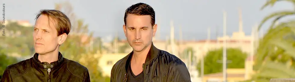 Zwei Männer in Jacken stehen vor einem Hintergrund mit Pflanzen und Gebäuden.